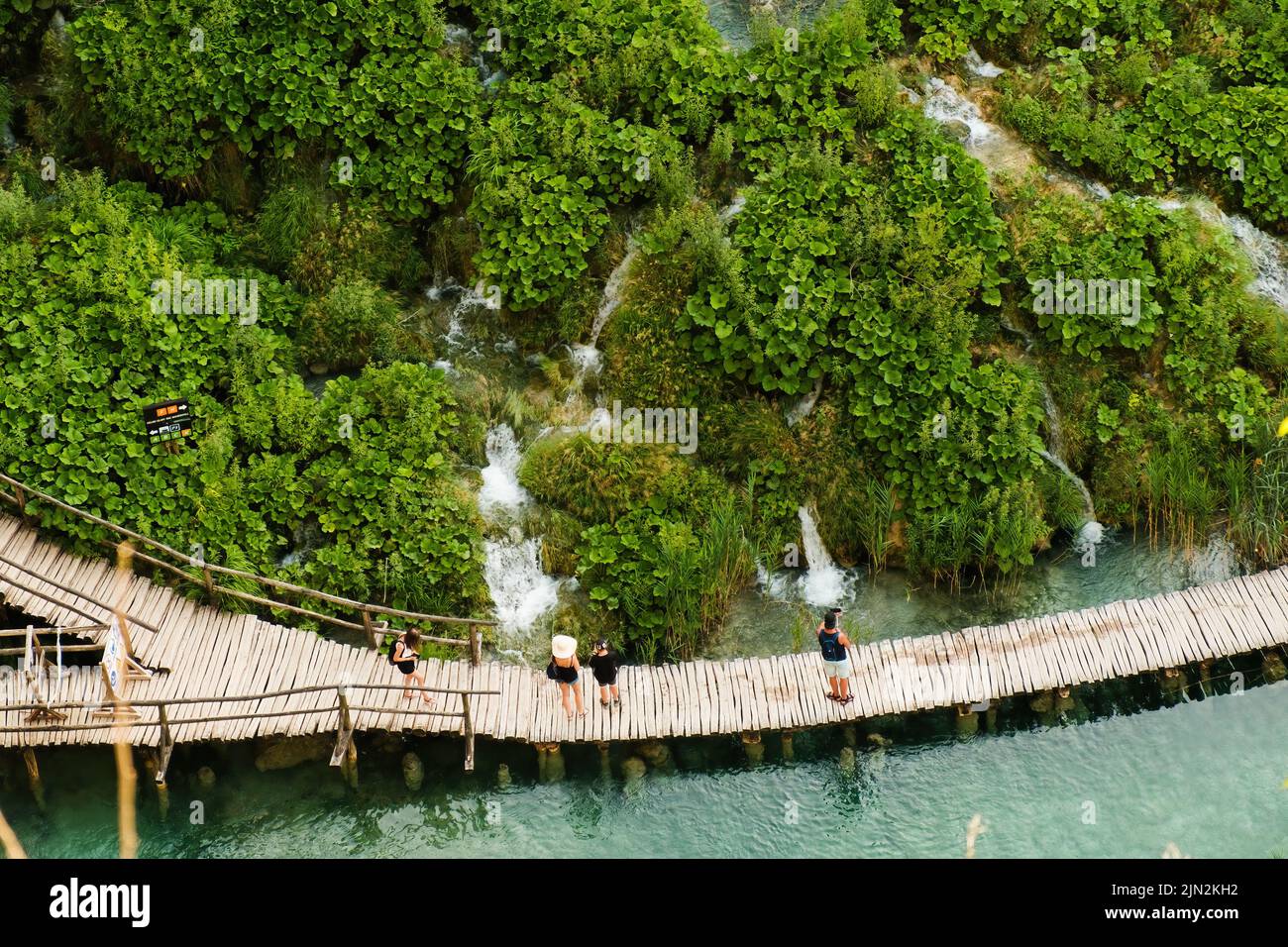 Wooden footbridge between aquamarine lake and hilly green bank. Tourists discover nature of famous national natural park reserve on Plitvice lakes upper view Stock Photo