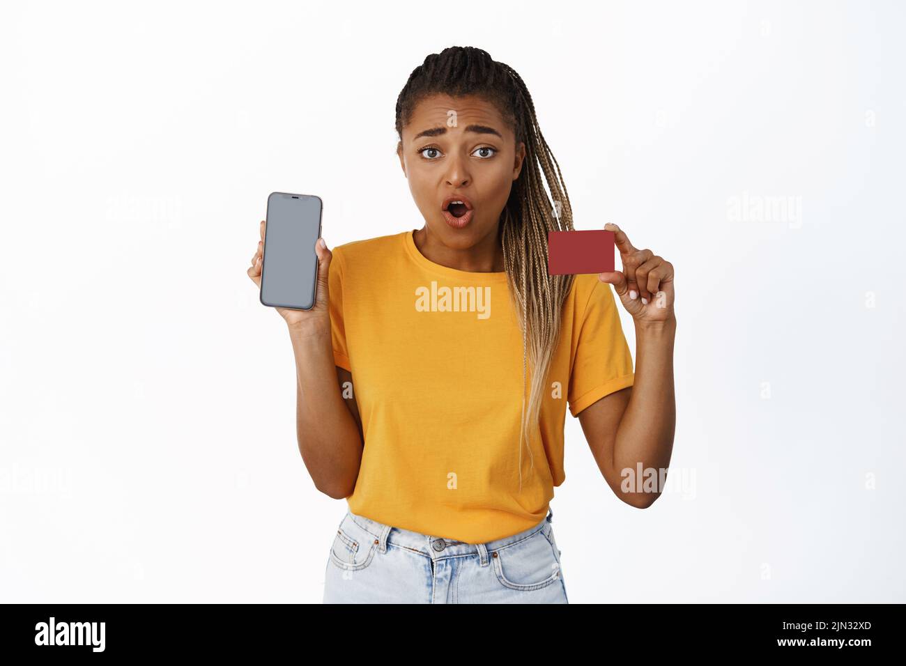 Surprised african american woman shows credit card and mobile phone screen, banking app or account balance on smartphone, looking amazed, white Stock Photo