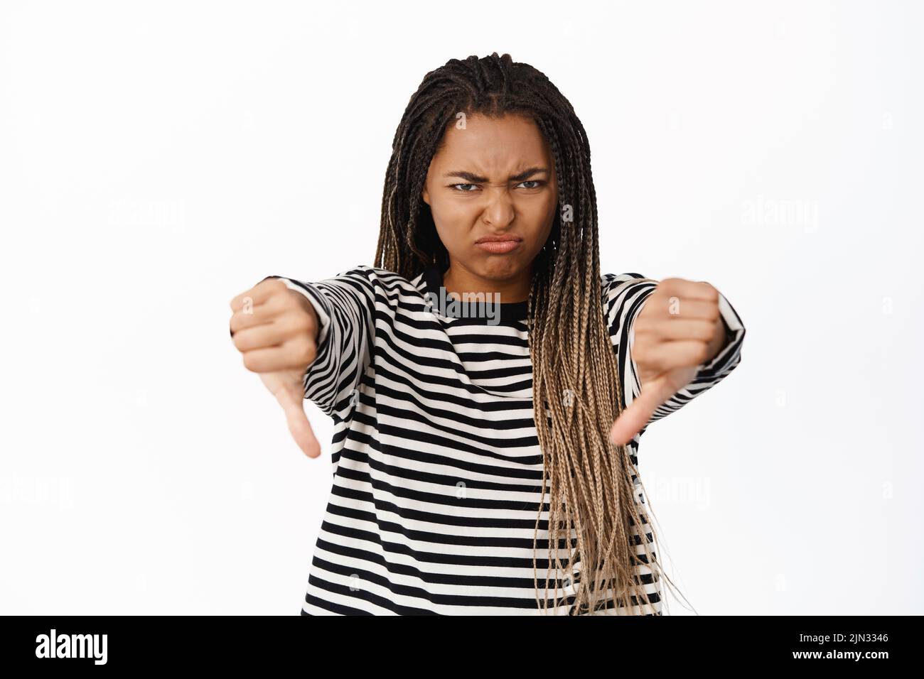 Portrait of disappointed black girl grimacing, showing thumbs down to express dislike and aversion, do not recommend smth, white background Stock Photo