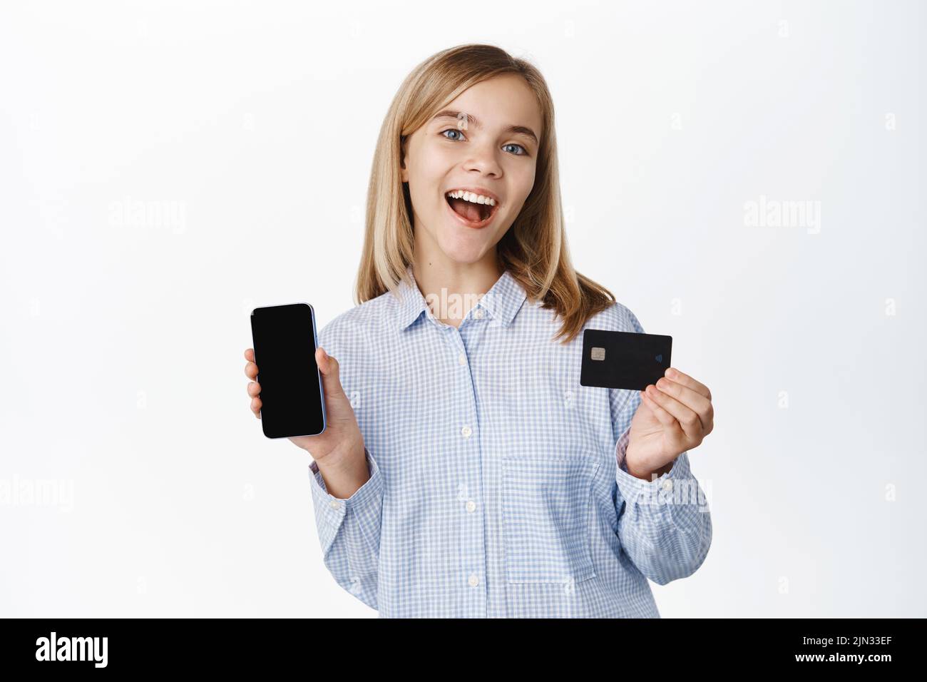 Happy smiling blond girl, teen child showing mobile phone screen and credit card, app interface, laughing excited, standing over white background Stock Photo