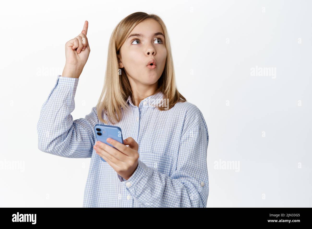Enthusiastic little girl, child in casual clothes, raising finger up, idea eureka gesture, using mobile phone app, smartphone application for kids Stock Photo