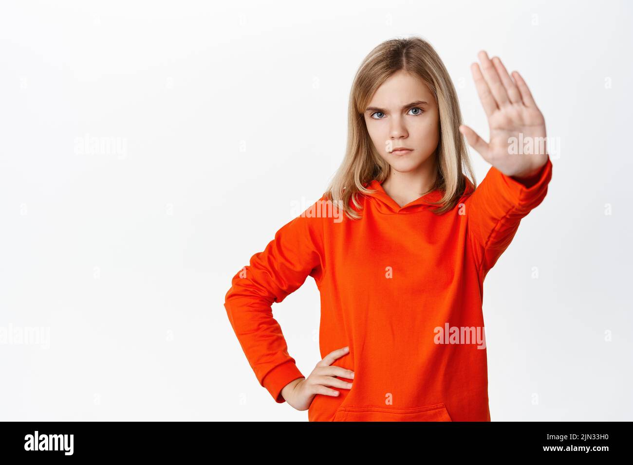 Stop. Little girl looks serious, extends one arm to say no, rejecting, prohibit action, forbid smth, standing in red hoodie over white background Stock Photo
