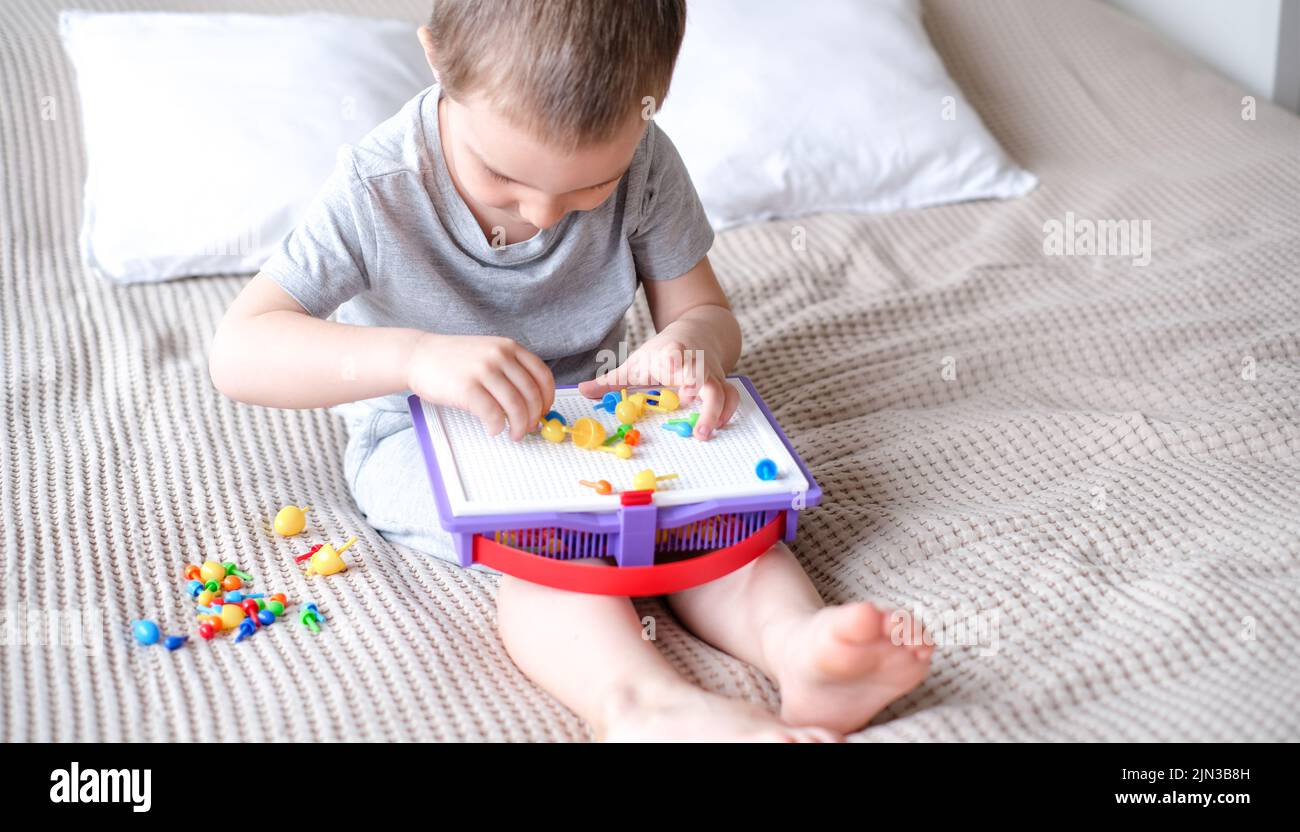 Little happy caucasian boy in a light room on the bed collects color mosaic to creating images Stock Photo