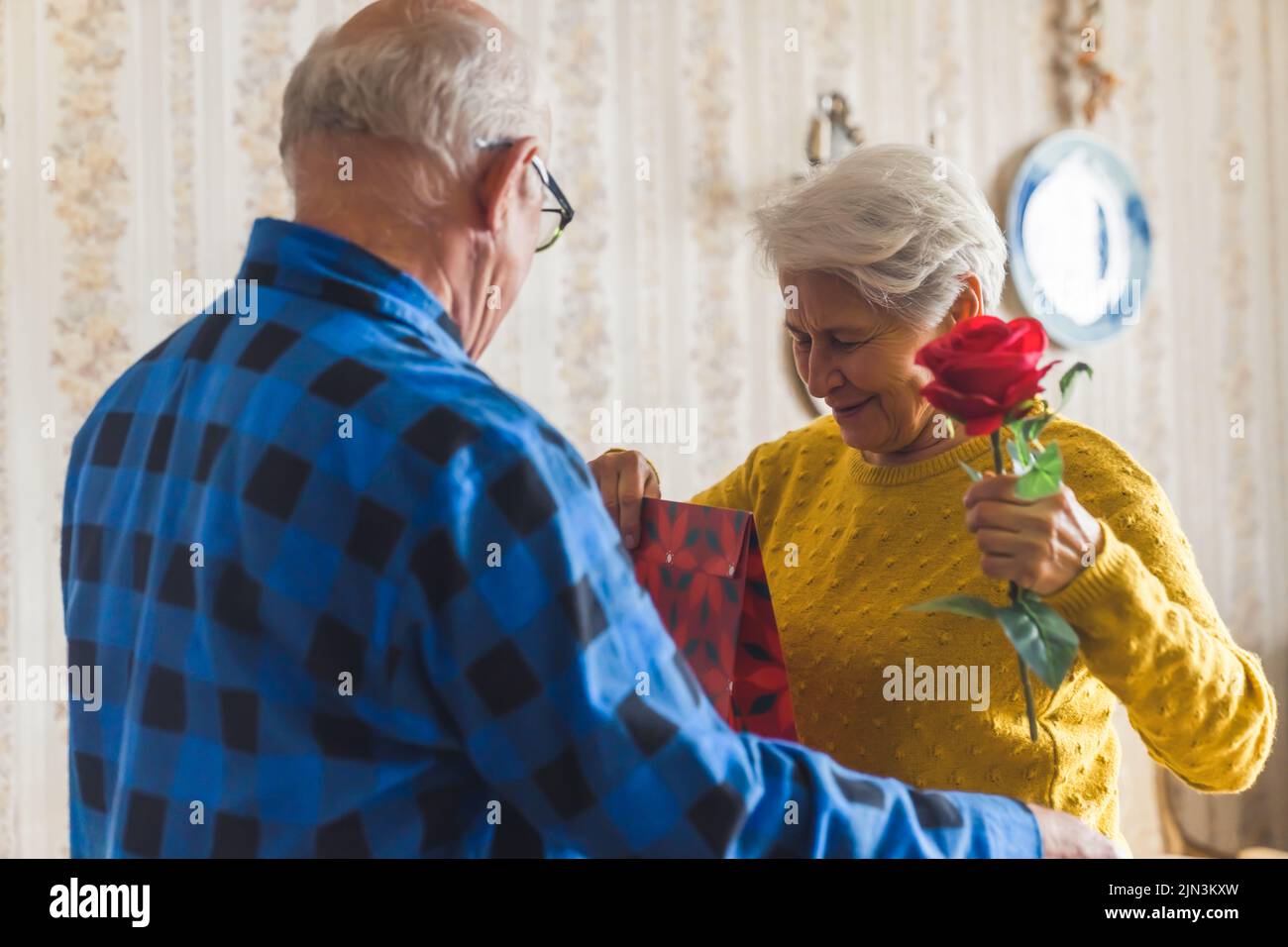Happy women's day. Caring and loving caucasian elderly husband surprising his wife with a red rose and a gift bag. High quality photo Stock Photo