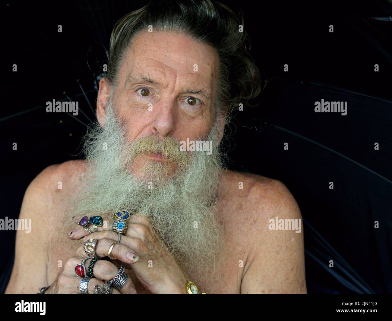 Surprised elderly eccentric Caucasian man with naked shoulders and grey beard holds a black umbrella with both his beringed hands. Stock Photo