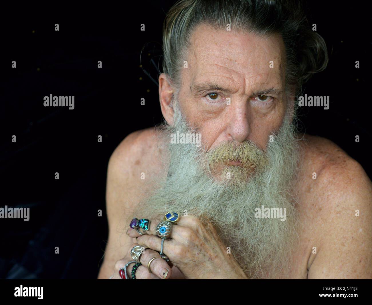 Worried wary elderly eccentric Caucasian man with naked shoulders and grey beard holds a black umbrella with both his beringed hands. Stock Photo