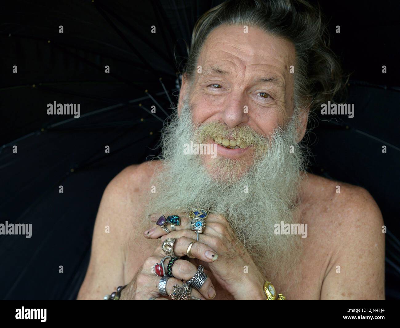 Smiling elderly eccentric Caucasian man with naked shoulders and grey beard holds a black umbrella with both his beringed hands. Stock Photo