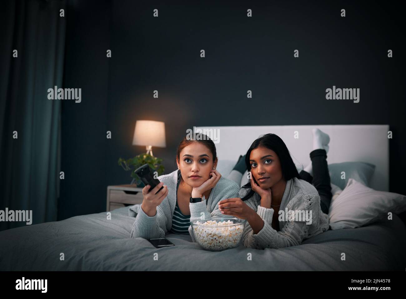 Lets see what we can watch. two young women eating popcorn while watching a movie at home. Stock Photo
