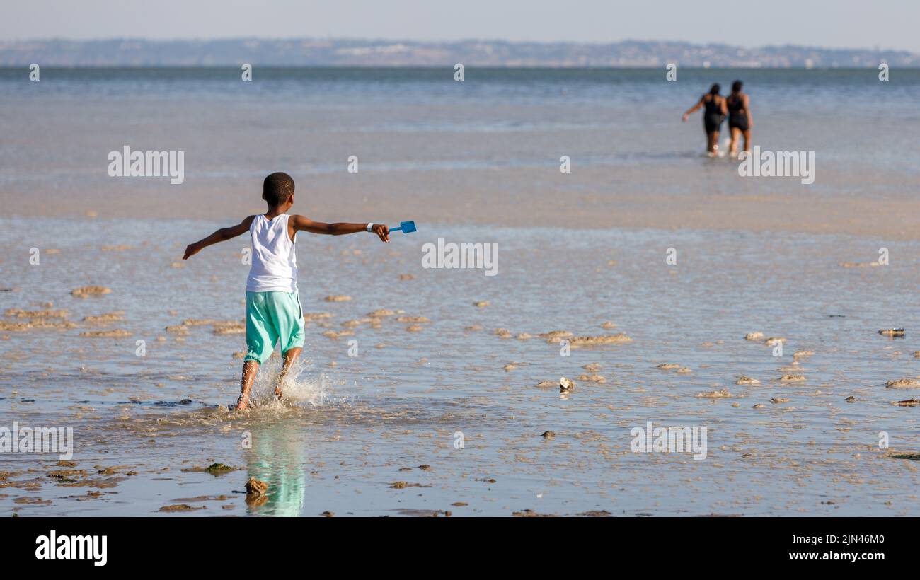 Heatwave UK. A young boy at the beach holding a toy spade runs in dirty water with algae foam spume caused by hot weather and pollutants. Stock Photo