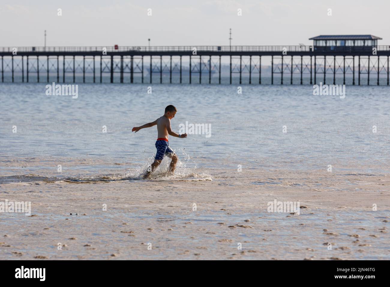 A boy runs in sea foam pollution at the beach with Southend pier in the background. Stock Photo