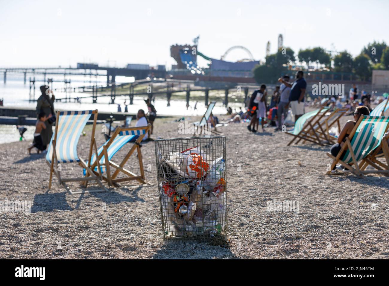 A trash can on a beach with deck chairs and people blurred in the background on a hot sunny day in summer. Rubbish bin. Stock Photo