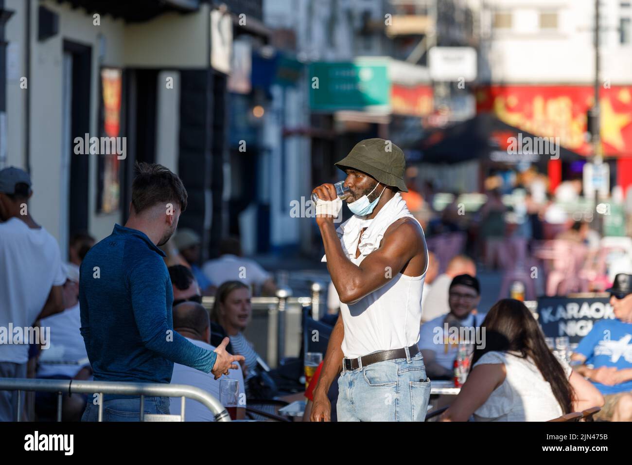 A black man wearing a bucket hat and covid mask drinks beer with his white friend in a pub garden during the English summer heatwave of 2022 Stock Photo