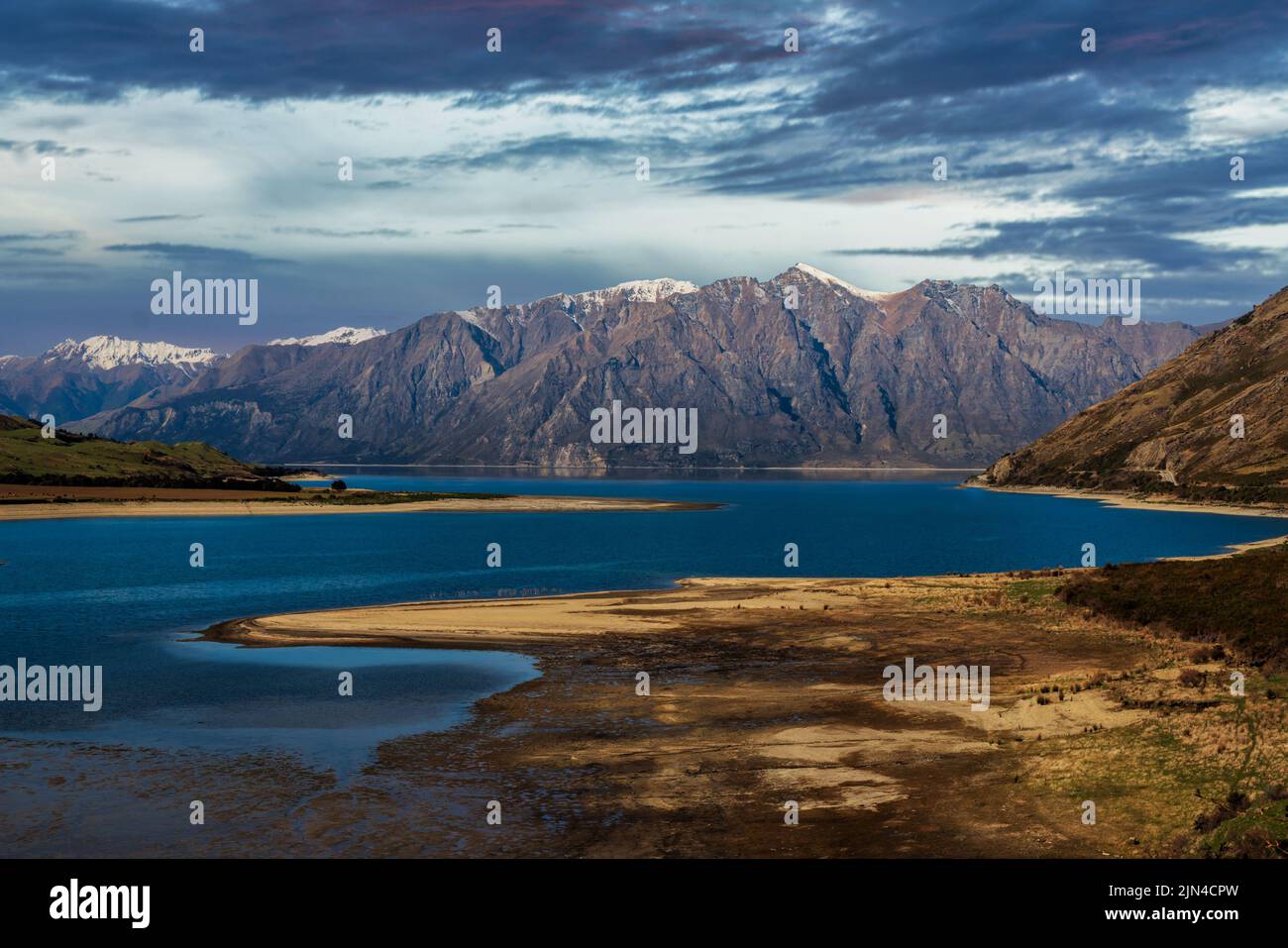 Lake Hawea foreshore with a dusting of snow on the mountain range peaks under a threatening cloudscape Stock Photo