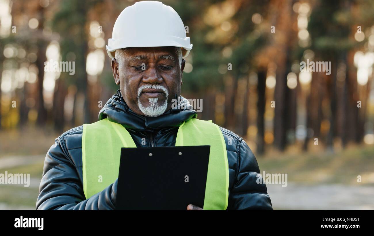 Mature concentrated forestry engineer technician forester in protective helmet stands in park assesses situation makes plan for cutting trees observes Stock Photo
