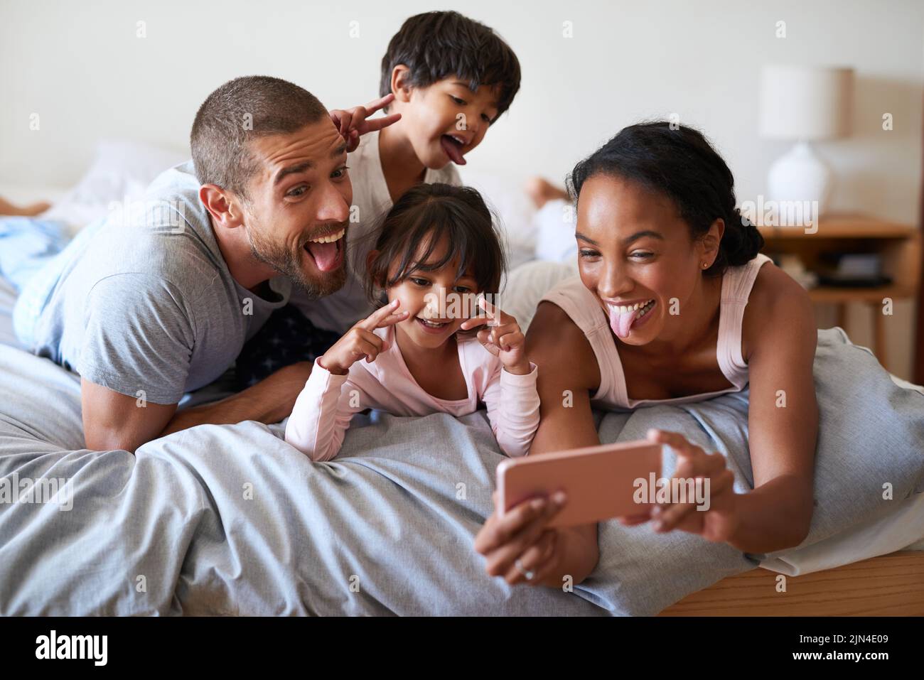 Silly faces and selfies in the morning. a beautiful young family of four taking a selfie with a cellphone in their bedroom at home. Stock Photo