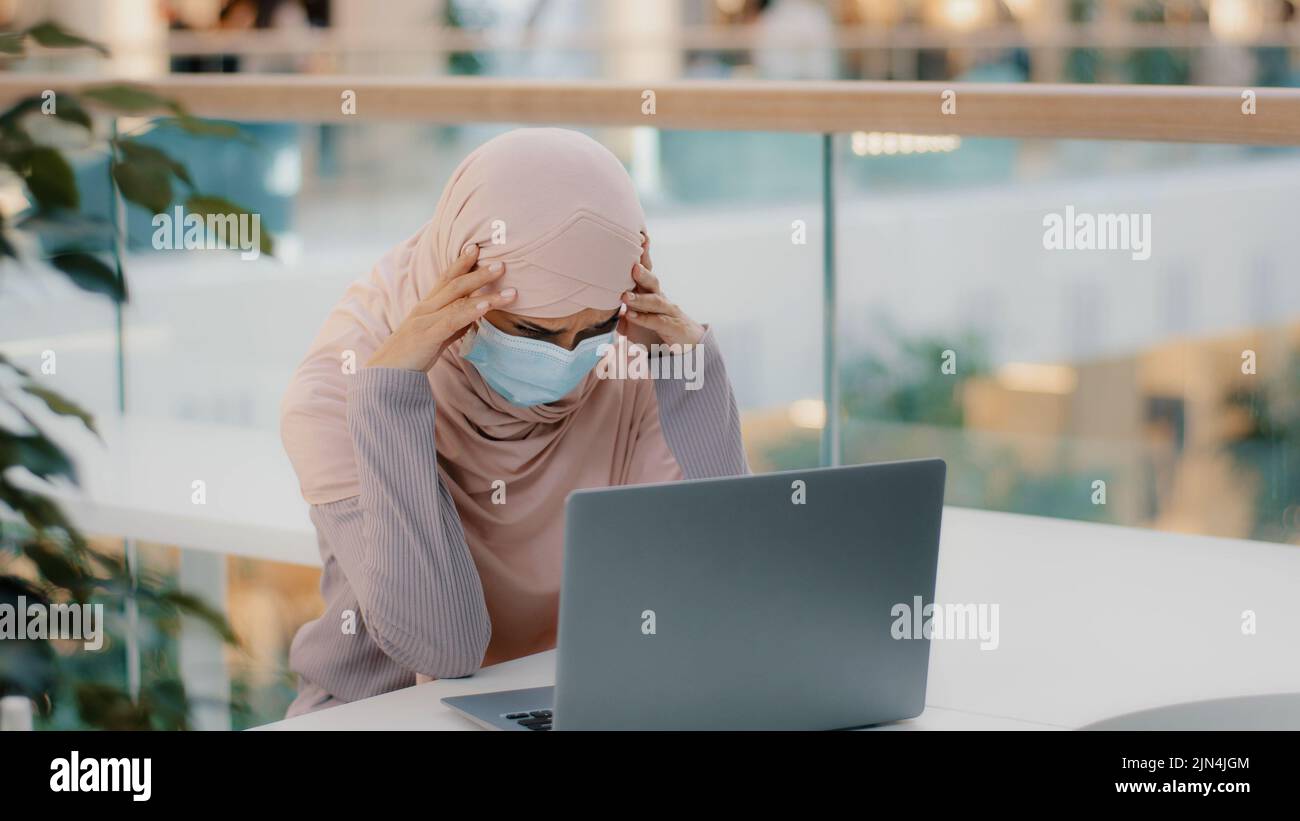 Pensive young muslim woman in medical mask in public place sitting typing on laptop arab girl in hijab writer journalist businesswoman student Stock Photo