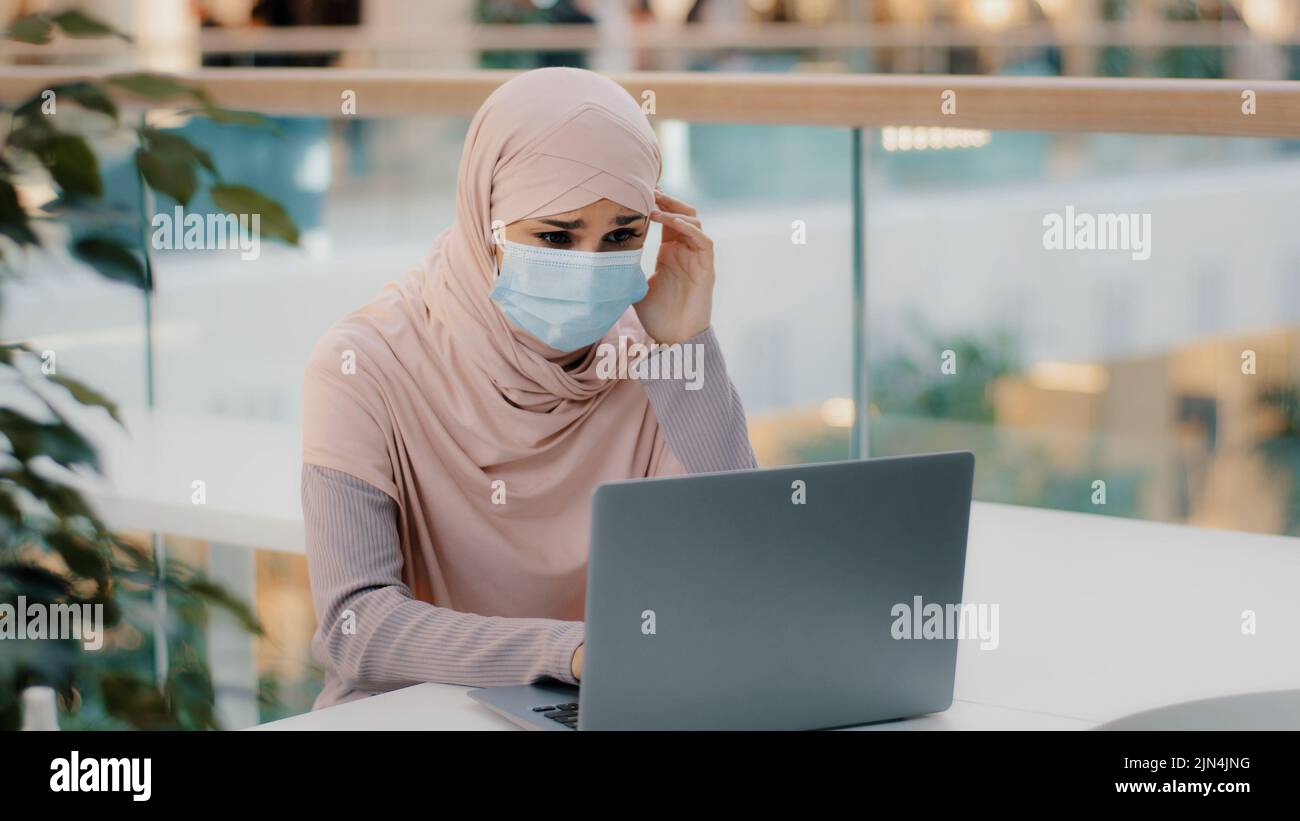 Pensive young muslim woman in medical mask in public place sitting typing on laptop arab girl in hijab writer journalist businesswoman student Stock Photo