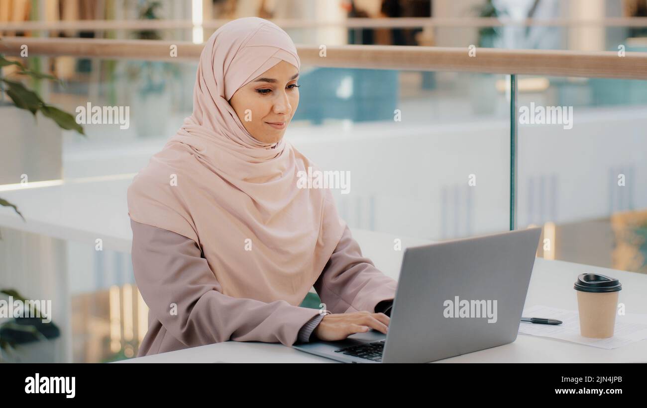 Happy young arab businesswoman sitting at office desk typing on laptop ...