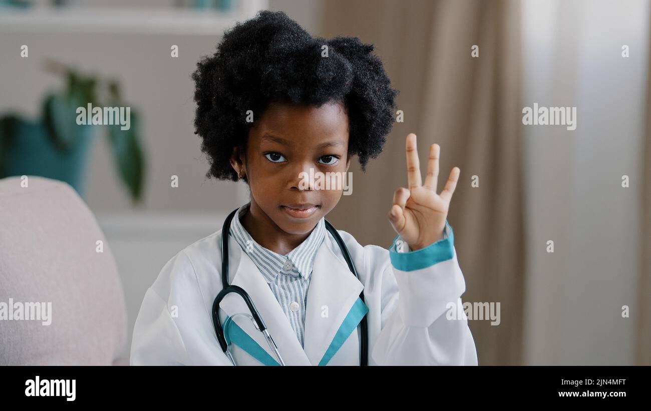 Close-up african american little girl standing in medical clothes playing pretending to be doctor charming child looking at camera smiling shows OK Stock Photo