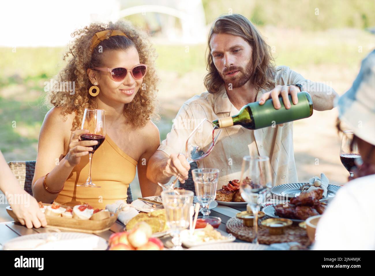 Portrait of young couple pouring red wine into glass while enjoying dinner party with friends outdoors in Summer Stock Photo