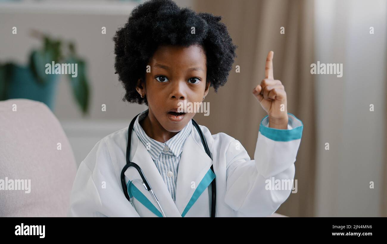 Close-up little cute pretty kid girl in medical clothes pretending to be doctor plays looking at camera posing raises index finger comes up with idea Stock Photo