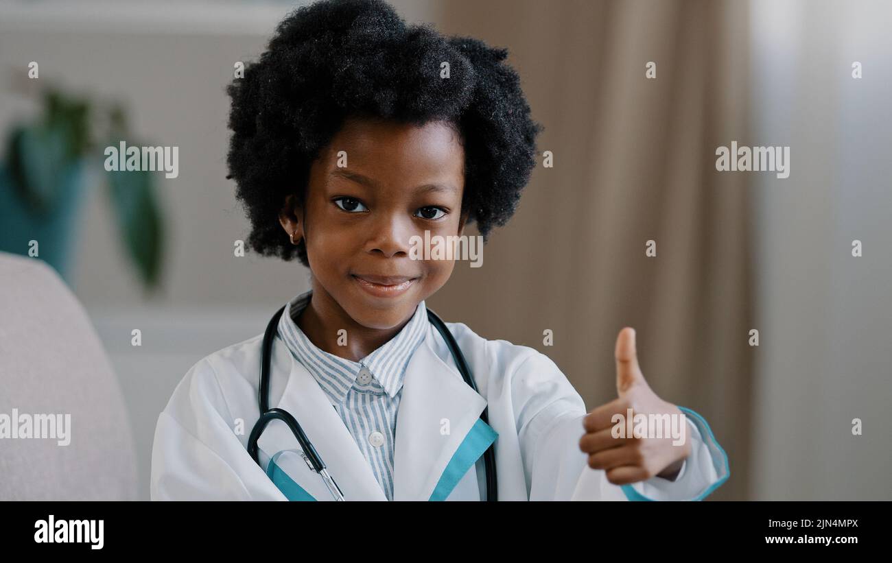 Close-up little cute kid girl in medical gown looking at camera posing indoors smiling pretending be doctor plays nurse showing thumb up gesture Stock Photo