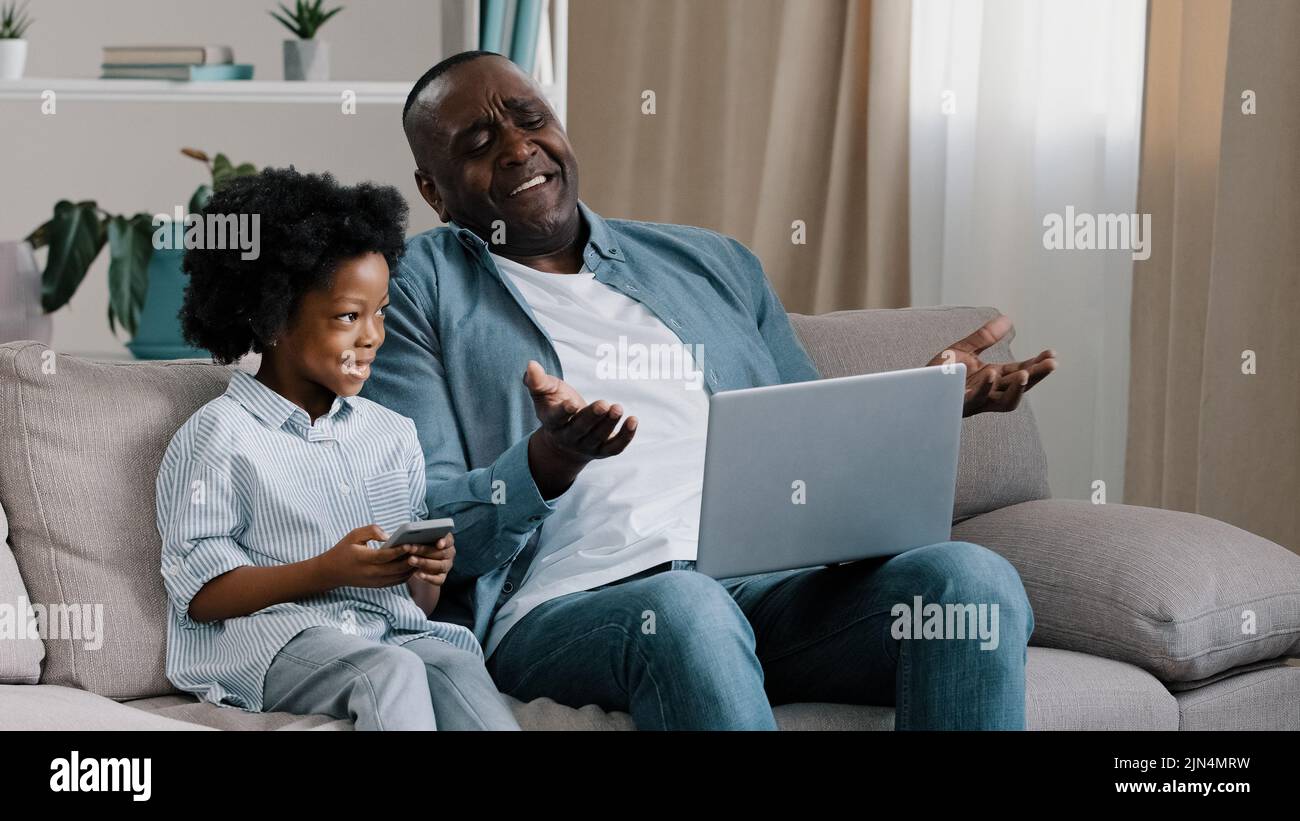 Upset male freelancer sitting on sofa in room with laptop dissatisfied with result loses online game gets bad news little cute daughter sitting next Stock Photo