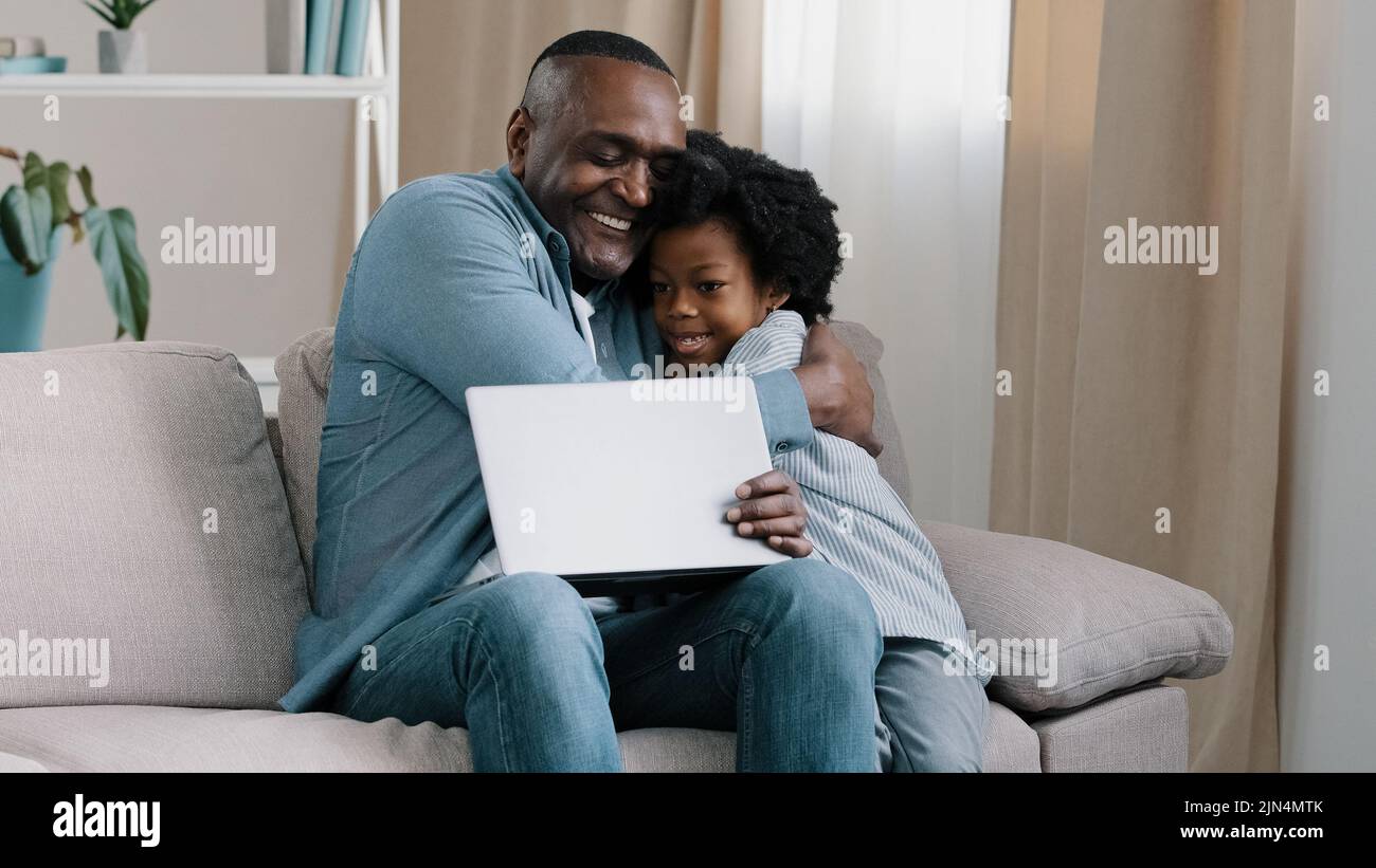 Adult african american male freelancer sitting on sofa working on laptop little daughter interferes with dad work asks for attention loving father Stock Photo