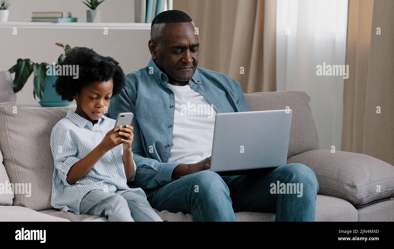 Upset male freelancer sitting on sofa in room with laptop dissatisfied with result loses online game gets bad news little cute daughter sitting next Stock Photo
