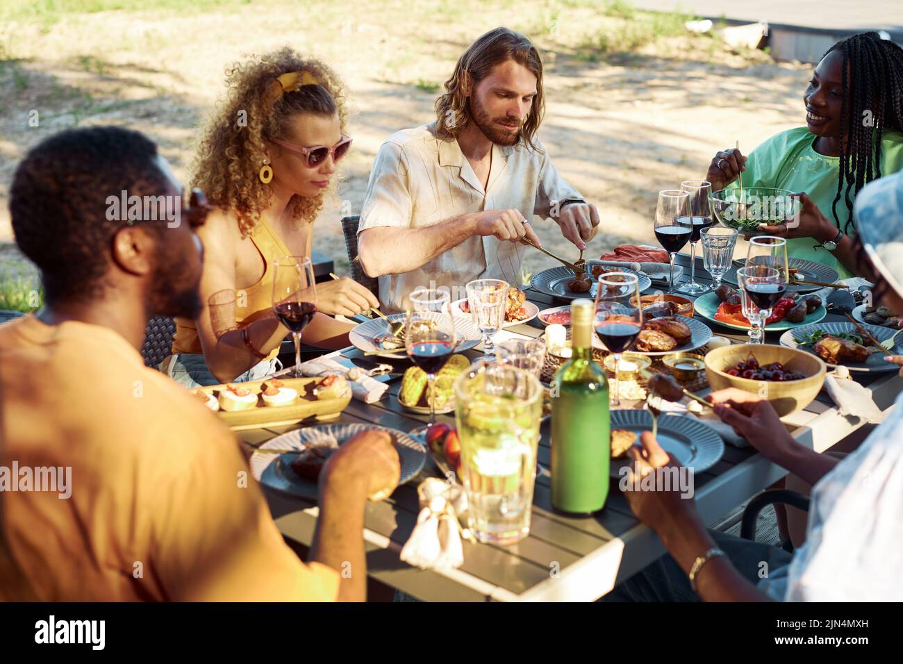 Diverse group of people enjoying dinner together outdoors in Summer sitting at table with delicious homemade feast Stock Photo