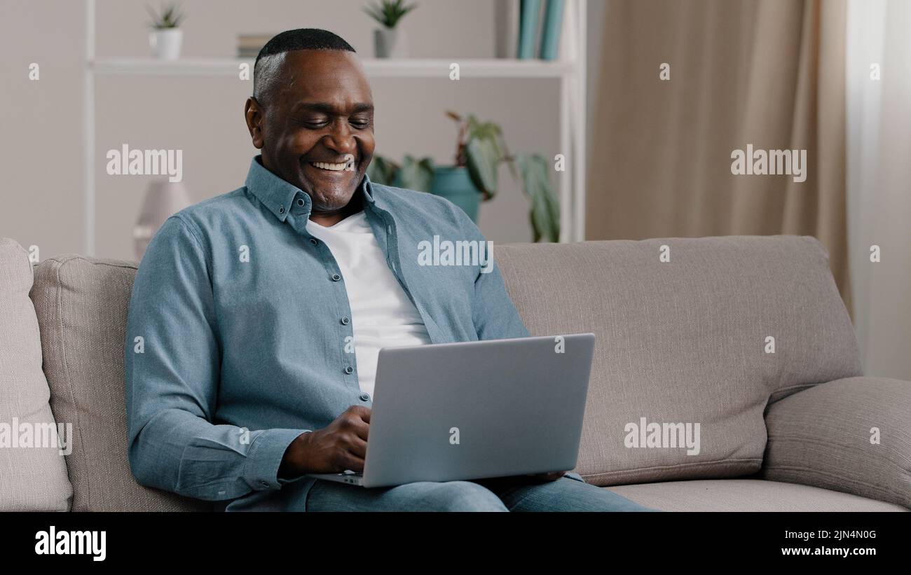 Mature happy african american man sitting on sofa at home relaxing smiling chatting on laptop via webcam talking on video call friendly conference Stock Photo