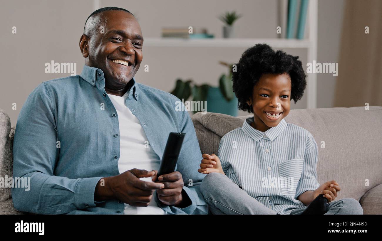 Close-up happy african american father and daughter laughing watching funny tv show movie relaxing on couch man holding remote control showing thumb Stock Photo