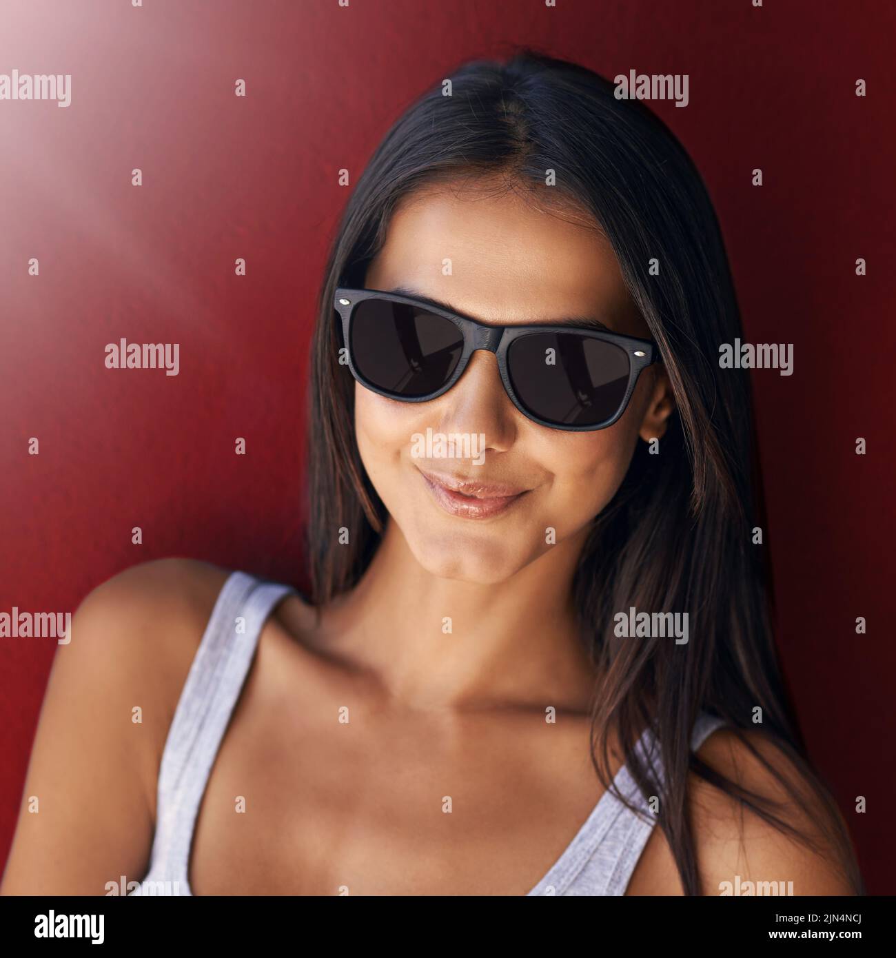 Cool, confident and beautiful woman wearing sunglasses and smiling against a red wall with lens flare. Face of a cheerful, stunning and happy female Stock Photo
