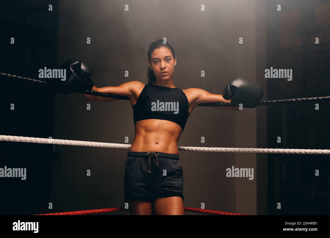 Muscular female athlete looking at the camera while leaning against the ropes of a boxing ring. Female boxer getting ready for a boxing match. Stock Photo