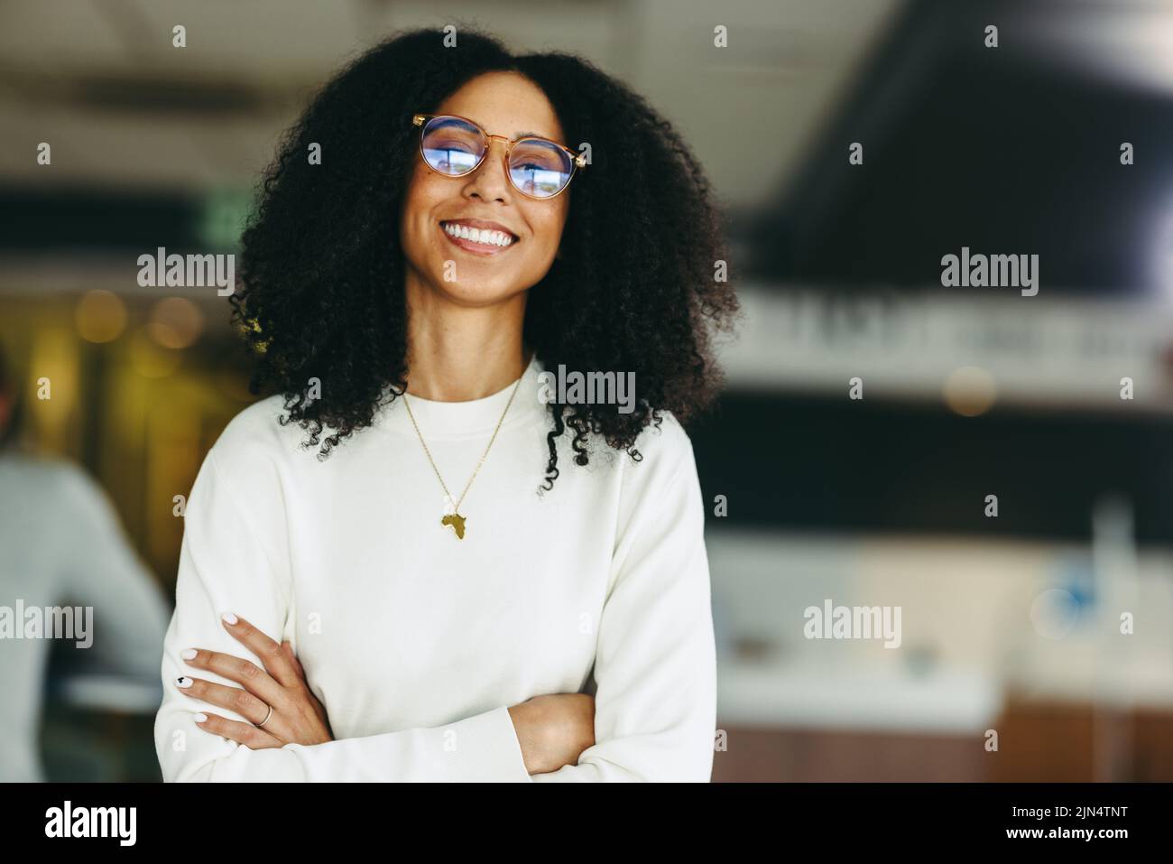 Confident young businesswoman smiling at the camera while standing with her arms crossed. Happy black businesswoman working remotely in a co-working s Stock Photo