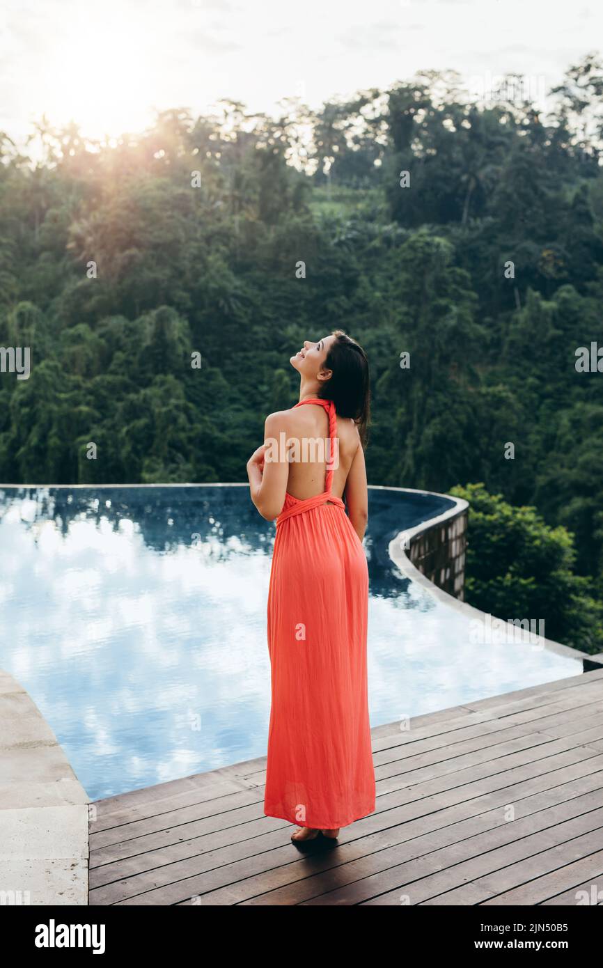 Rear view of young female model standing on the edge of the pool and looking away. Caucasian woman enjoying her vacation at luxury resort. Stock Photo