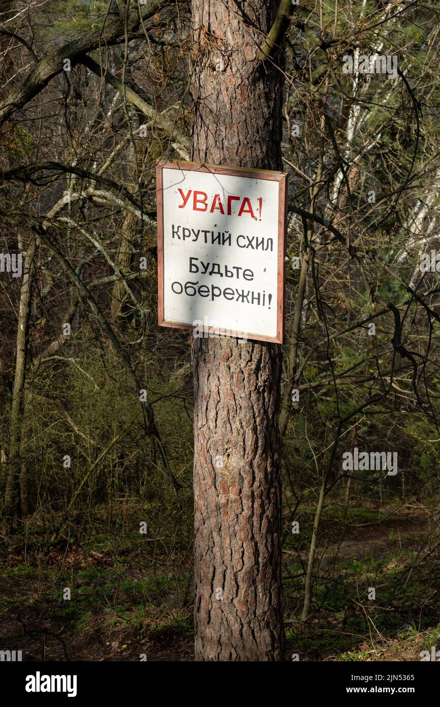 White sign board with warning text on pine tree trunk in sunny spring forest. Translation from ukrainian: Warning! Steep slope Be careful! Stock Photo