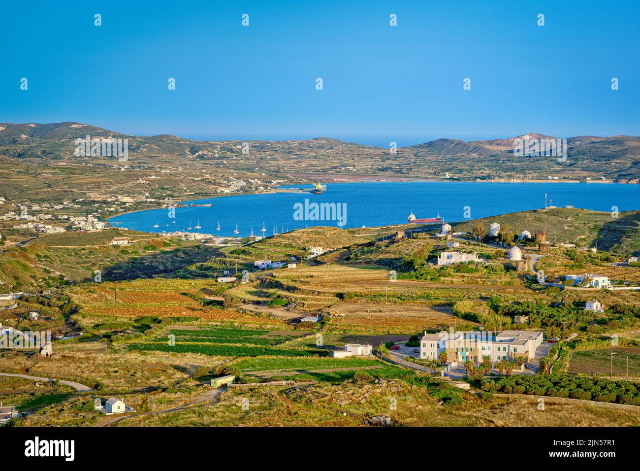 Beautiful view of Adamantas town, bay and villages of Milos island at sunset, Greece. Whitewashed houses, green hills, low sun, blue sea, clear sky, Stock Photo