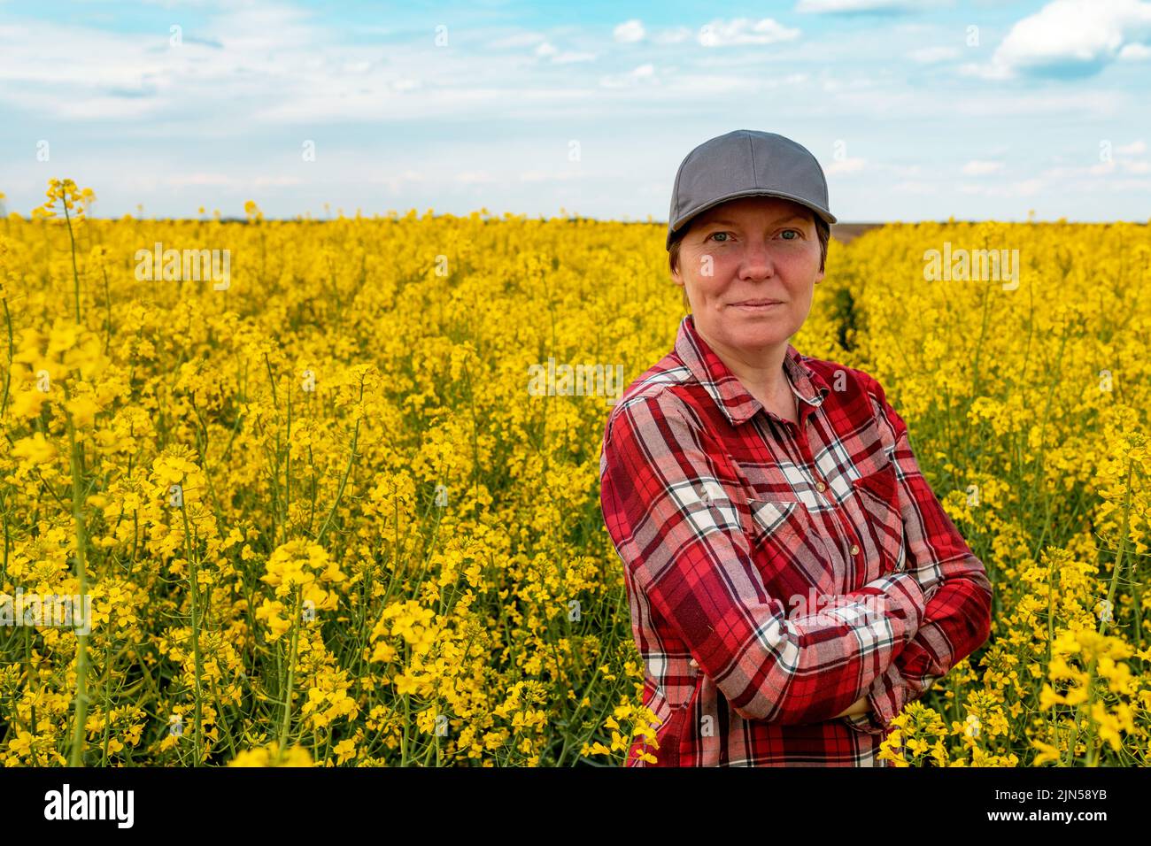 Confident and self-assured farm worker wearing red plaid shirt and trucker's hat standing in cultivated rapeseed field in bloom and looking over crops Stock Photo