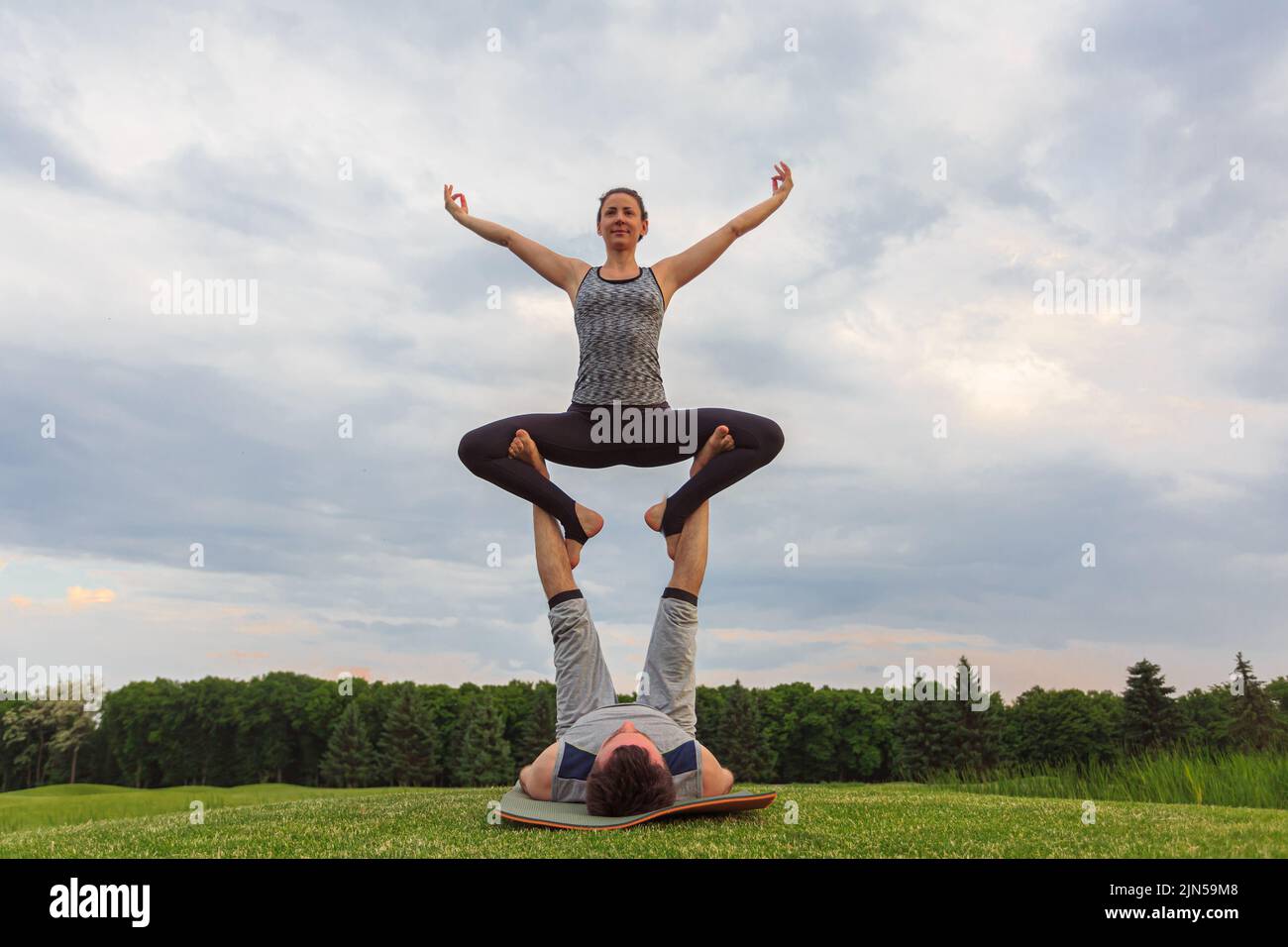 Young couple doing acro yoga in park. Man lying on grass and balancing woman in his feet Stock Photo