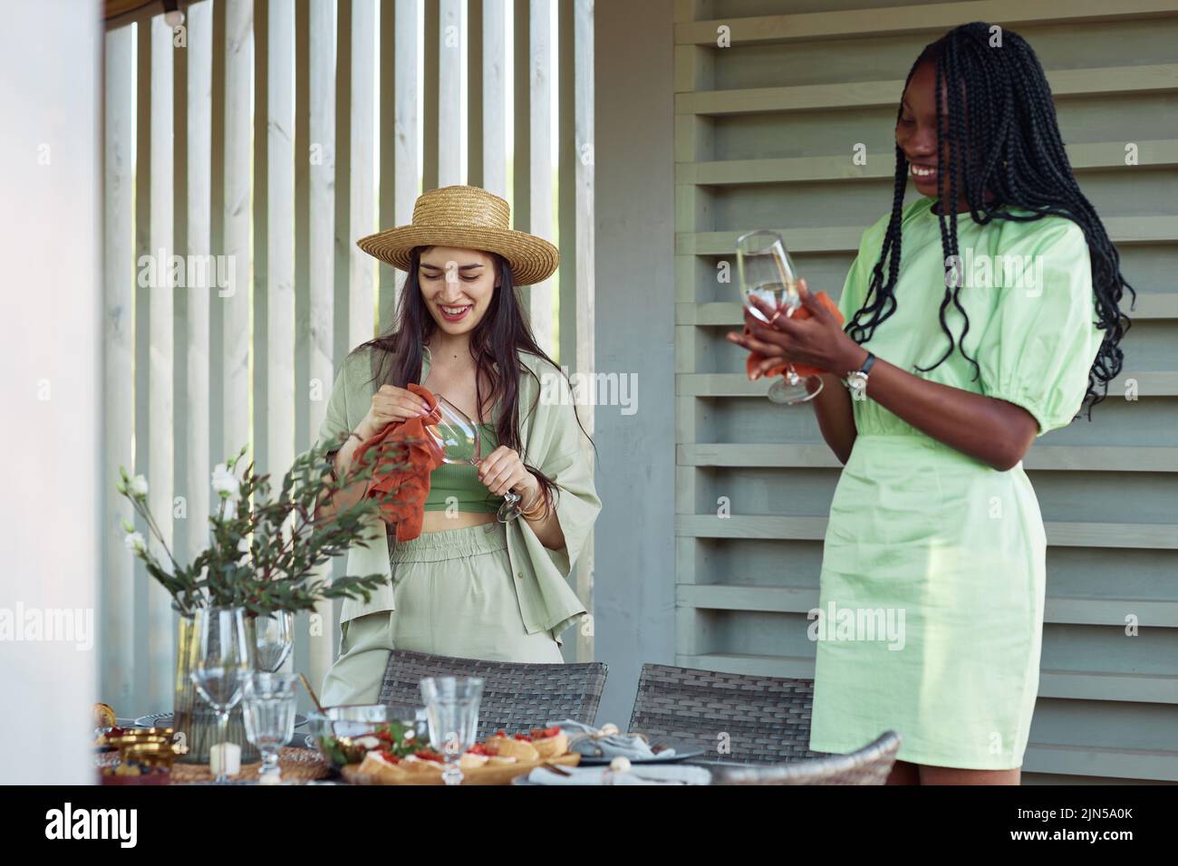 Waist up portrait of two young women setting up dinner table for party with friends outdoors in muted green color Stock Photo