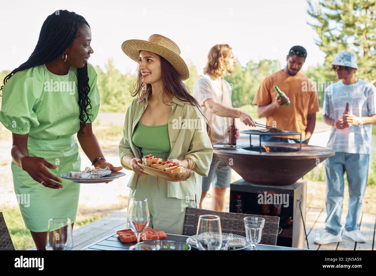 Diverse group of young people enjoying barbeque party outdoors in Sumer, focus on two smiling women serving food, copy space Stock Photo