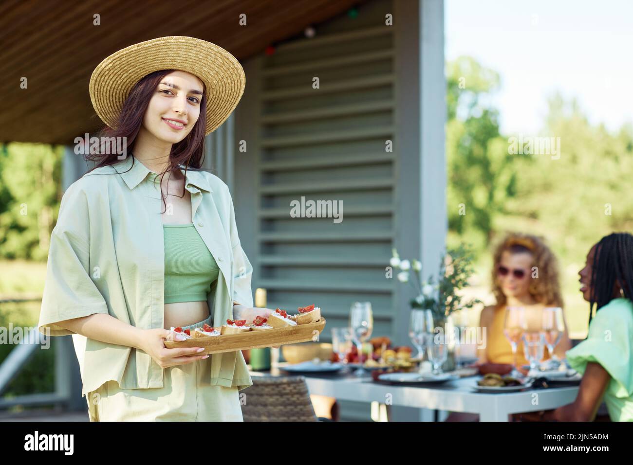 Waist up portrait of young woman holding tray of snacks looking at camera while enjoying outdoor party with friends in Summer, copy space Stock Photo