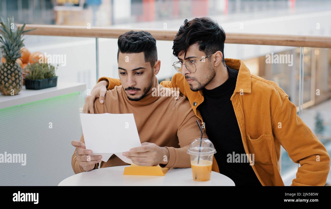 Two friends students sitting in cafe young worried man receiving letter reading bad news upset feels sad failing exam dismissal financial problem loss Stock Photo