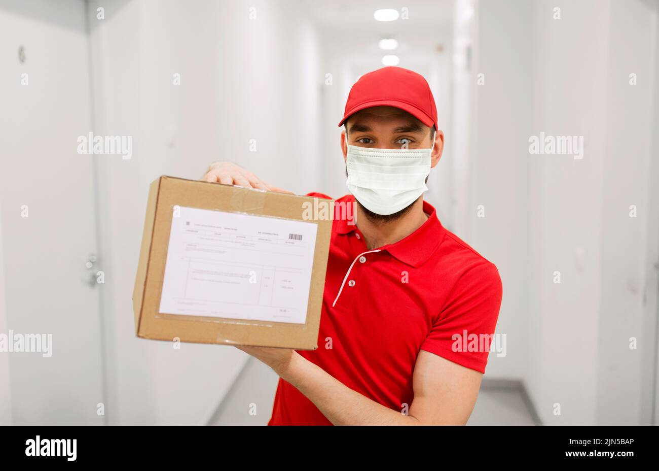 delivery man in mask with parcel box in corridor Stock Photo