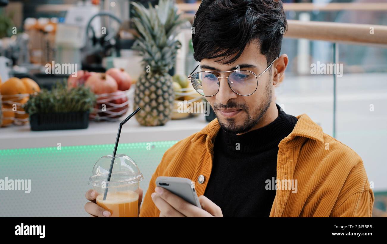 Young happy guy sits in cafe drinks freshly squeezed juice satisfied man holding mobile phone typing message email communicate with friends remote in Stock Photo