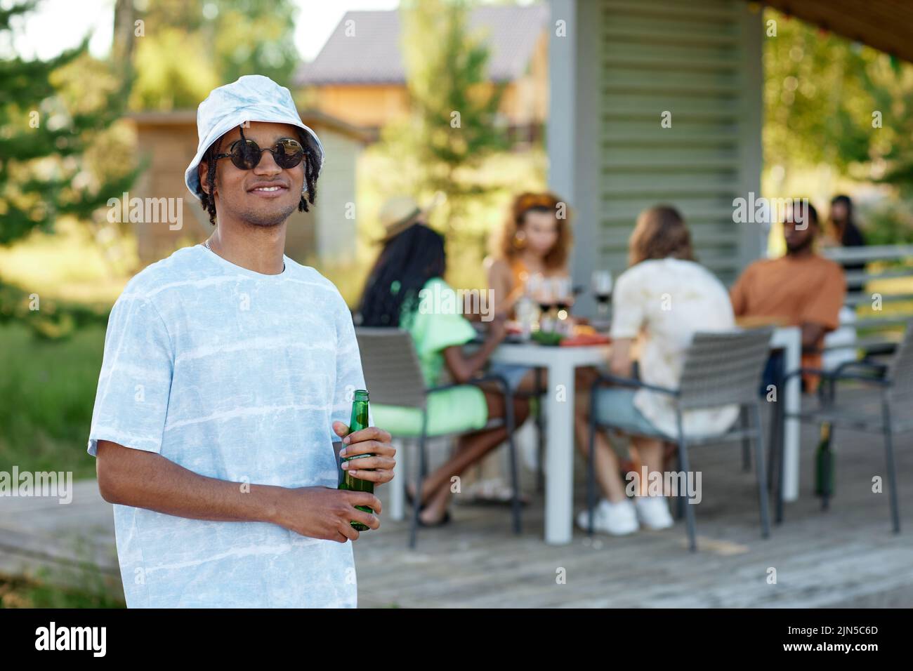 Waist up portrait of black young man wearing sunglasses smiling at camera and holding drink during outdoor party with friends in Summer, copy space Stock Photo