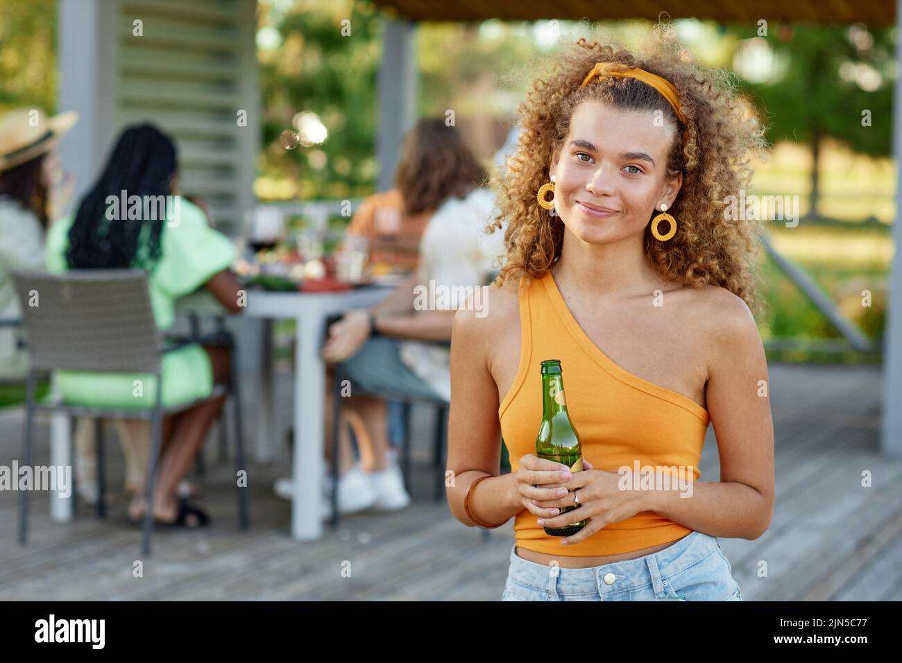 Waist up portrait of curly haired young woman smiling at camera and holding drink during outdoor party with friends in Summer, copy space Stock Photo