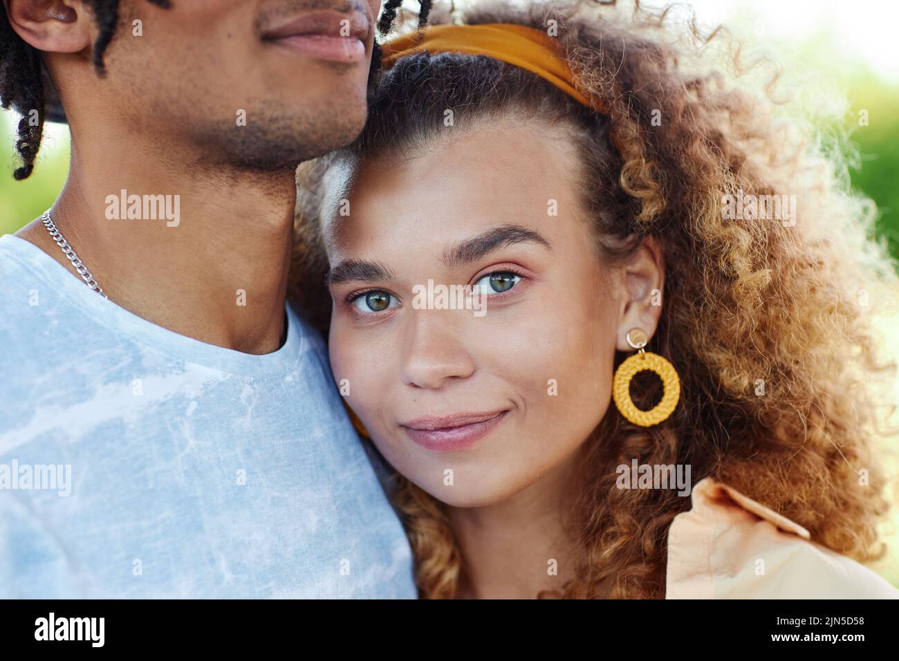 Close up portrait of curly hair young woman smiling at camera and embracing anonymous boyfriend during romantic date outdoors Stock Photo
