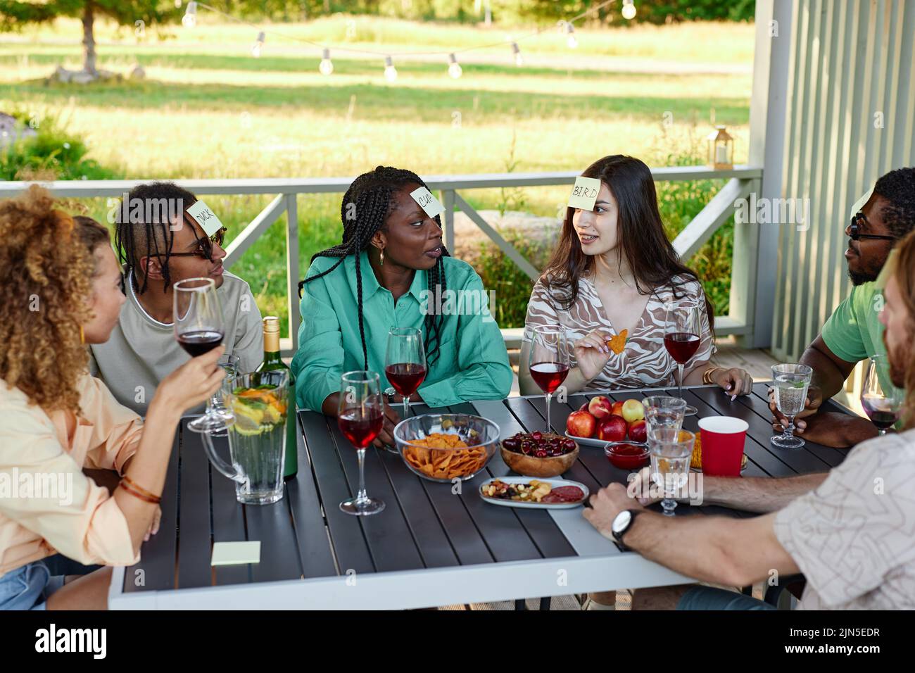 Diverse group of friends playing Guess who game while sitting at table outdoors in Summer Stock Photo